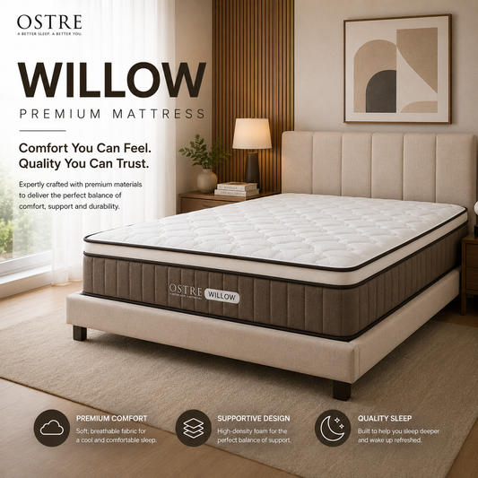 A staged bedroom scene showing an Ostre Willow premium mattress on a upholstered bed frame. The room includes a window with sheer curtains, a bedside table and lamp, wall art, and floor mats.